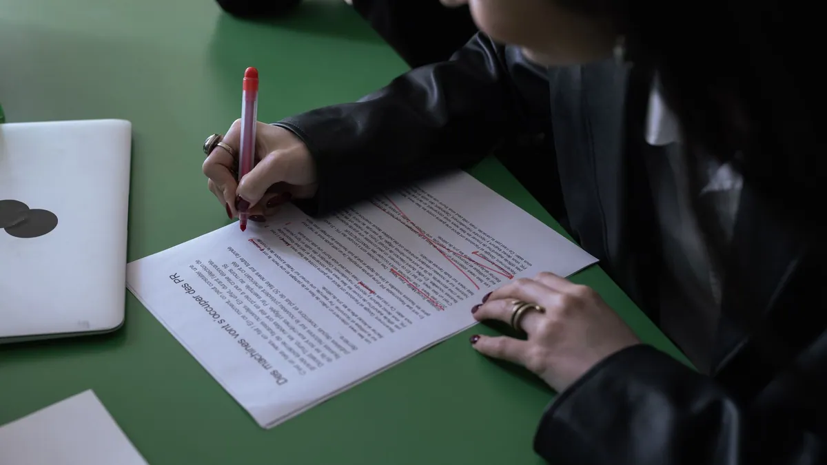 A translator post-editing a translation draft scored by a QE model, correcting it by hand with a red pen beside a laptop