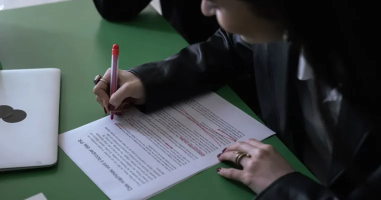 Woman in suit proofreads a document with a red pen, laptop nearby, embodying translation and proofre