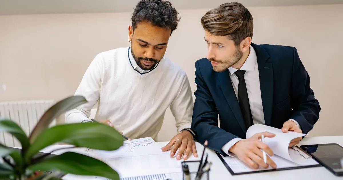 Two men analyze charts on documents, discussing MTPE cost savings.