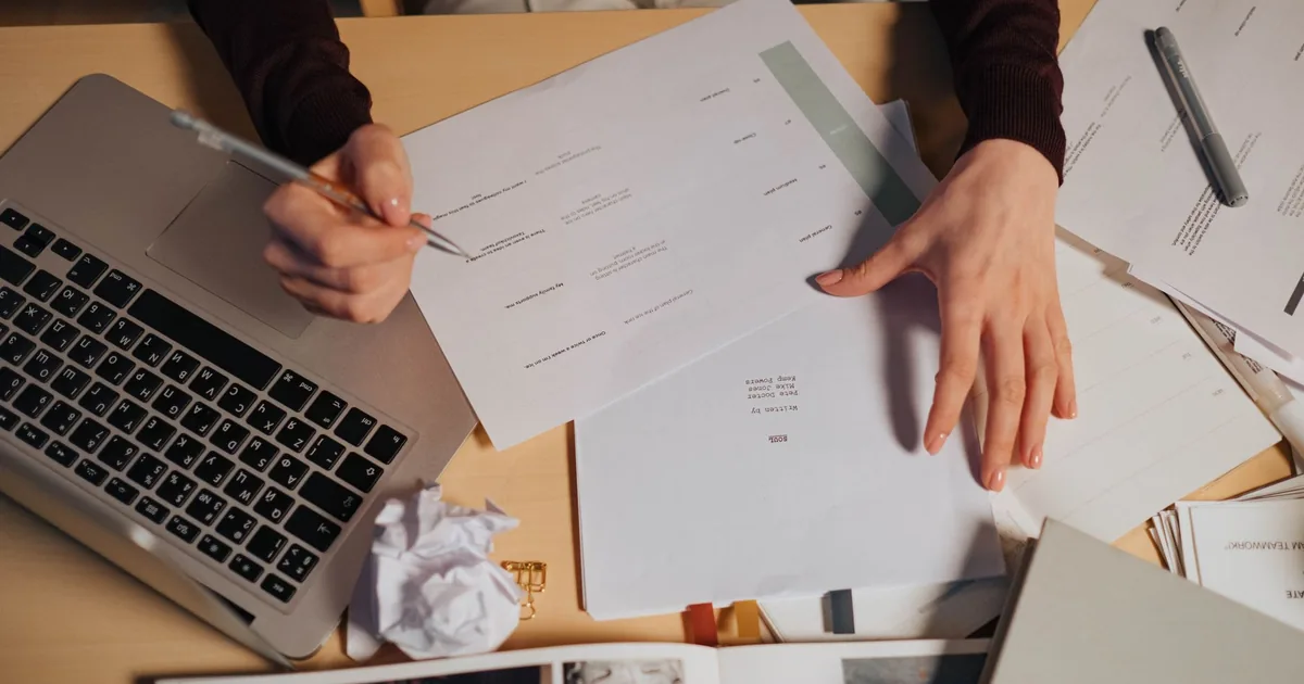 A person taking notes and working on a laptop surrounded by documents on a desk. - FAS Localize
